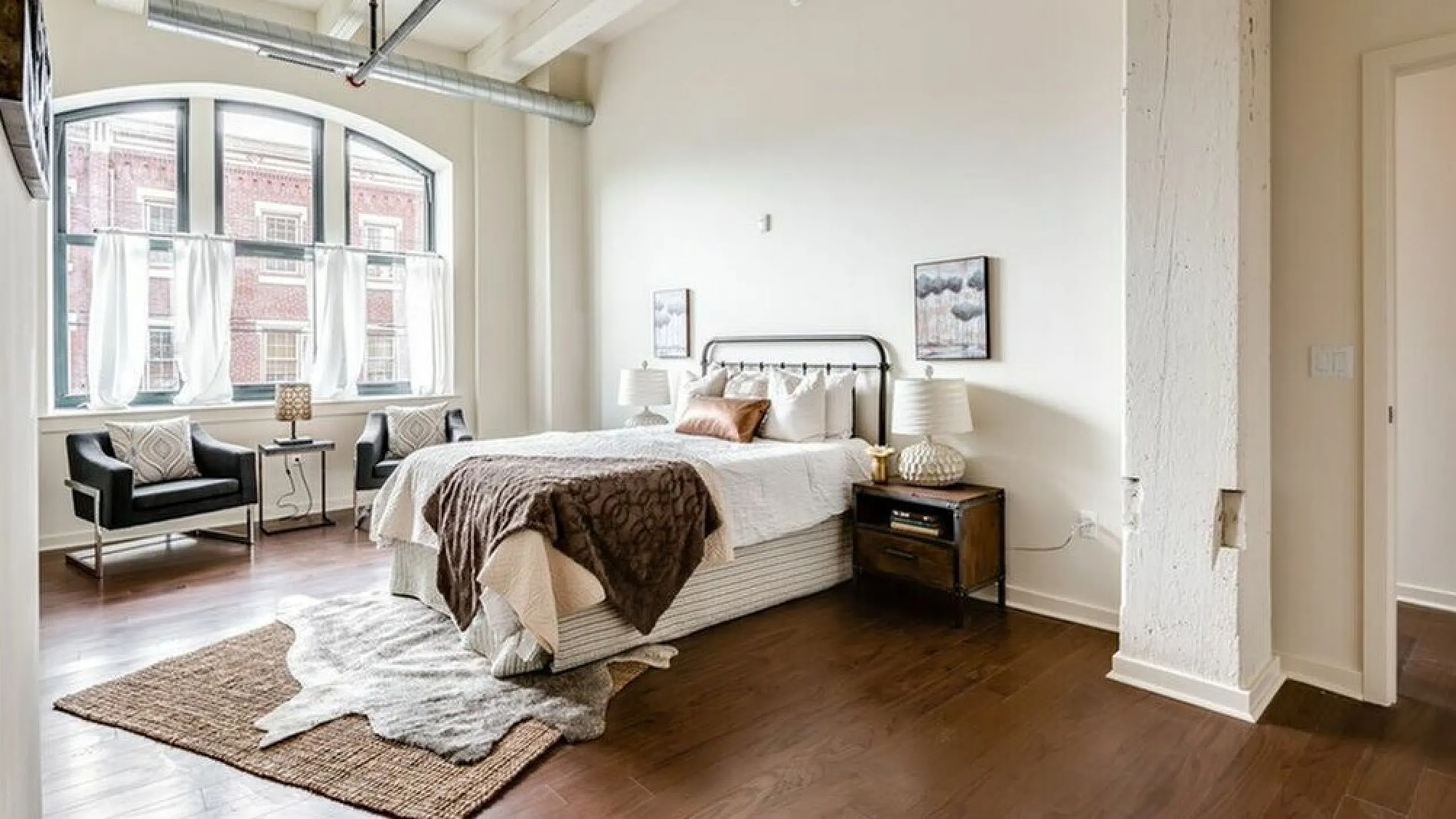 Modern loft bedroom with elegant decor, large industrial windows, and rich hardwood floors.