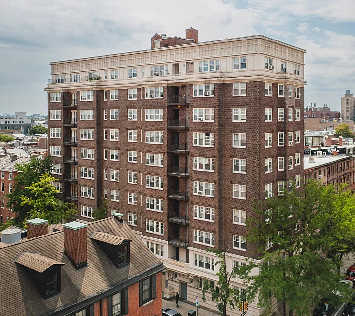 Charming red-brick apartment building nestled in a vibrant urban neighborhood with lush greenery and a distant cityscape view.