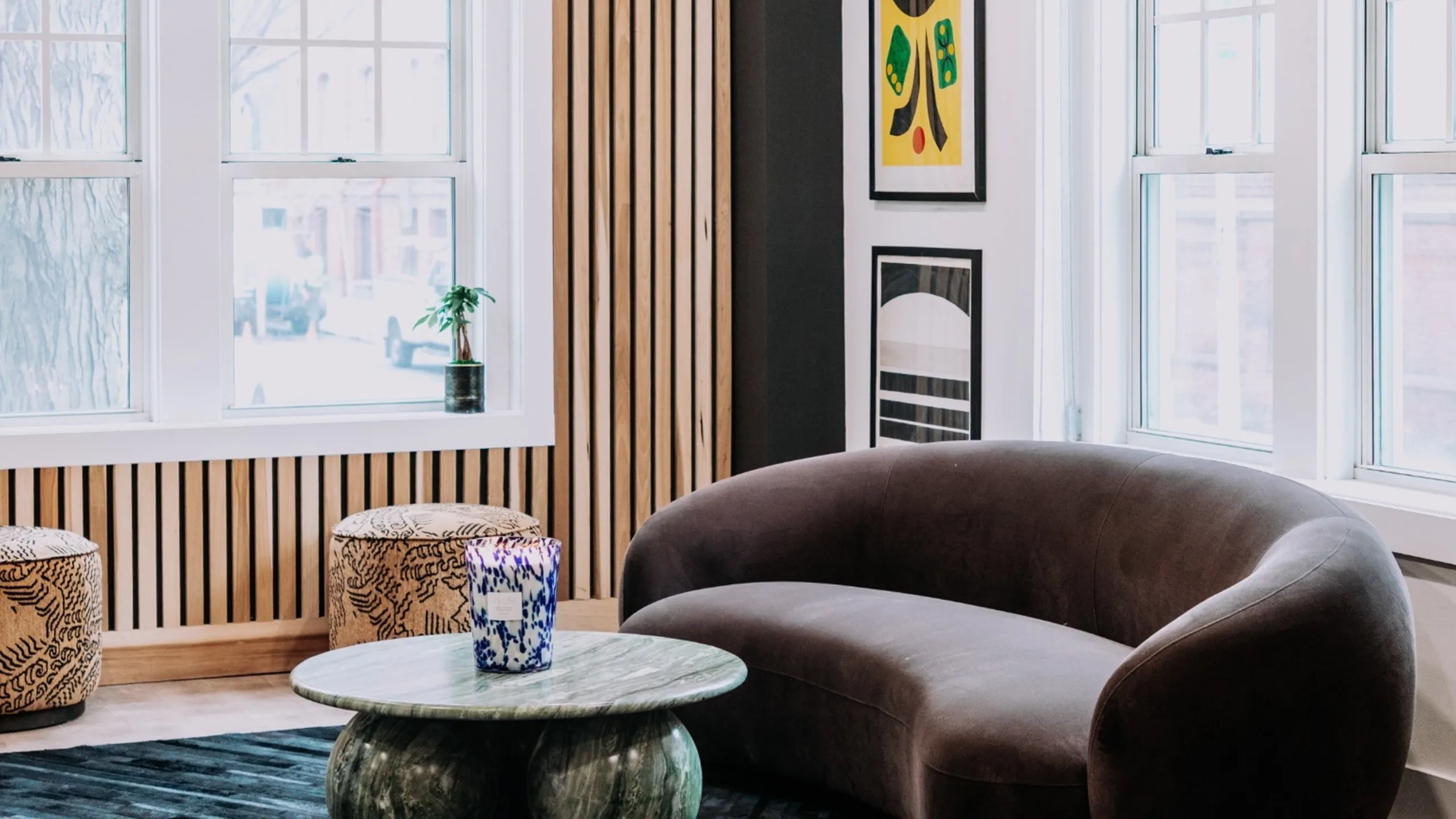 Chic and modern living space with a cozy brown sofa, stylish round marble table, and vibrant art decor. Large windows and natural wood accents create a bright and inviting atmosphere.