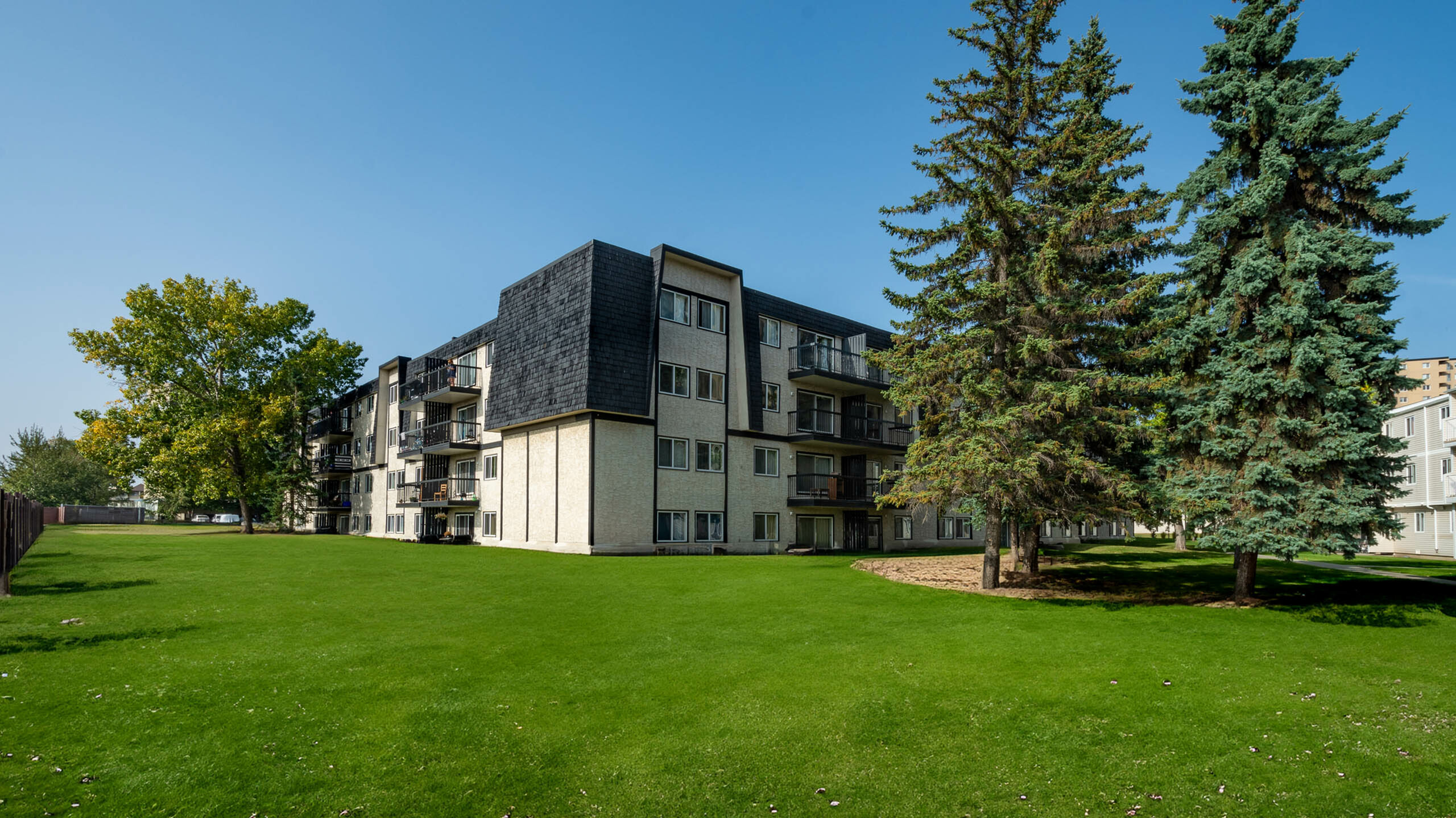 Charming apartment building surrounded by lush green lawns and tall trees under a clear blue sky.