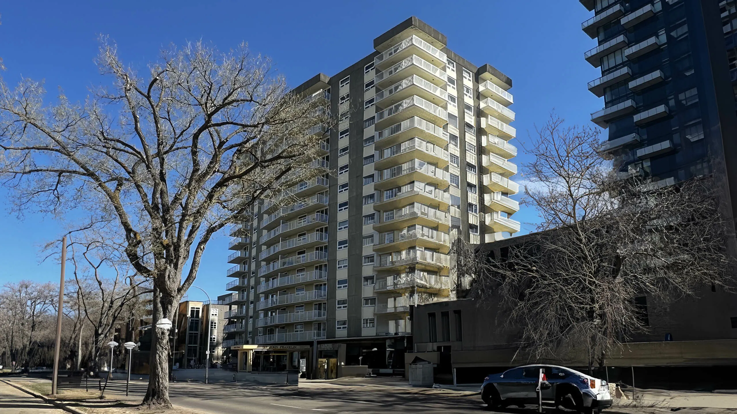 Modern apartment building with spacious balconies, set against a clear blue sky, offering urban living amidst scenic trees.
