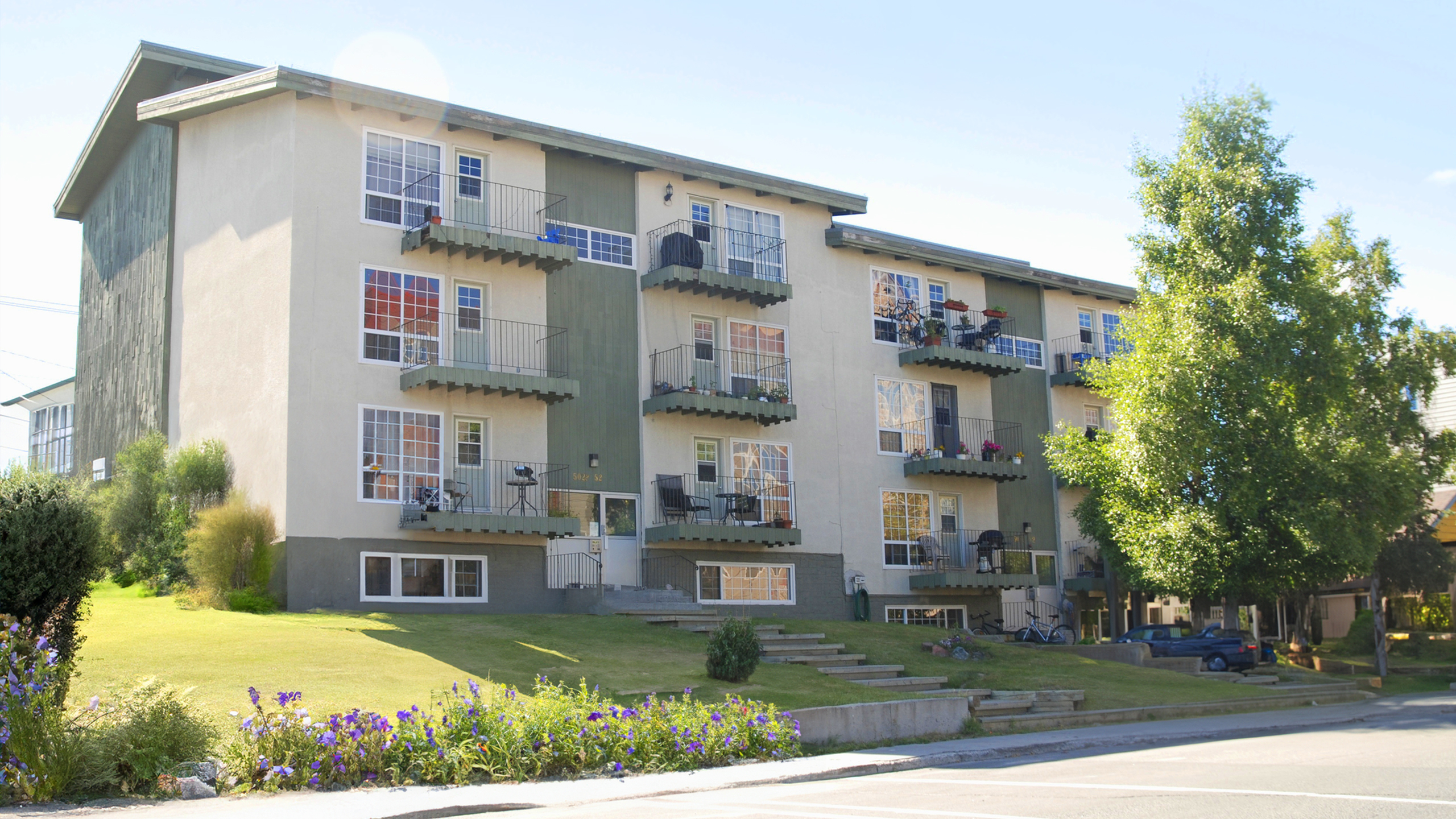 Charming three-story apartment building with cozy balconies, surrounded by lush greenery and vibrant flowers, capturing modern living in a serene neighborhood.