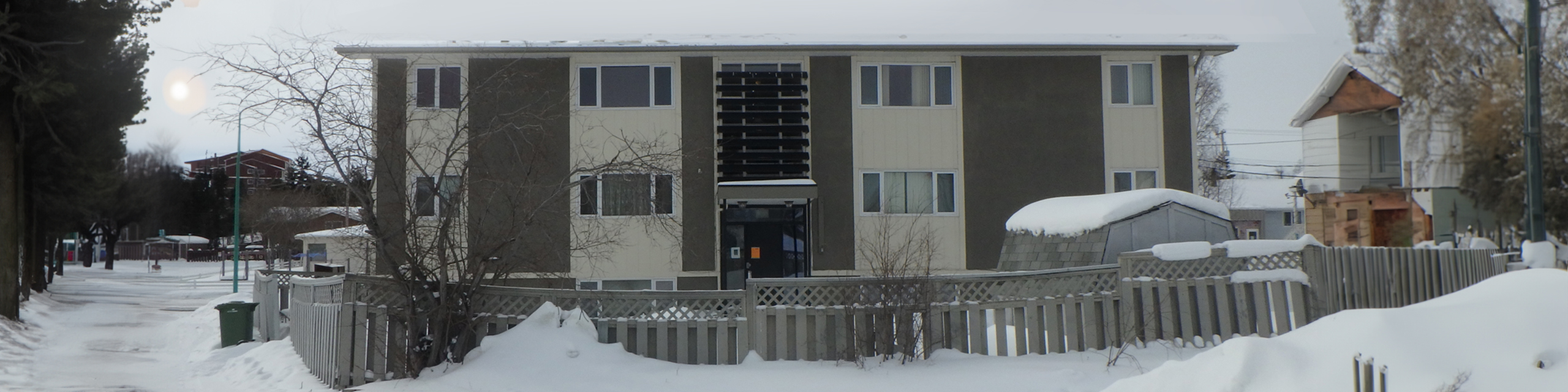 Charming two-story apartment building in a snowy neighborhood, featuring large windows and a fenced yard, perfect for cozy winter living.