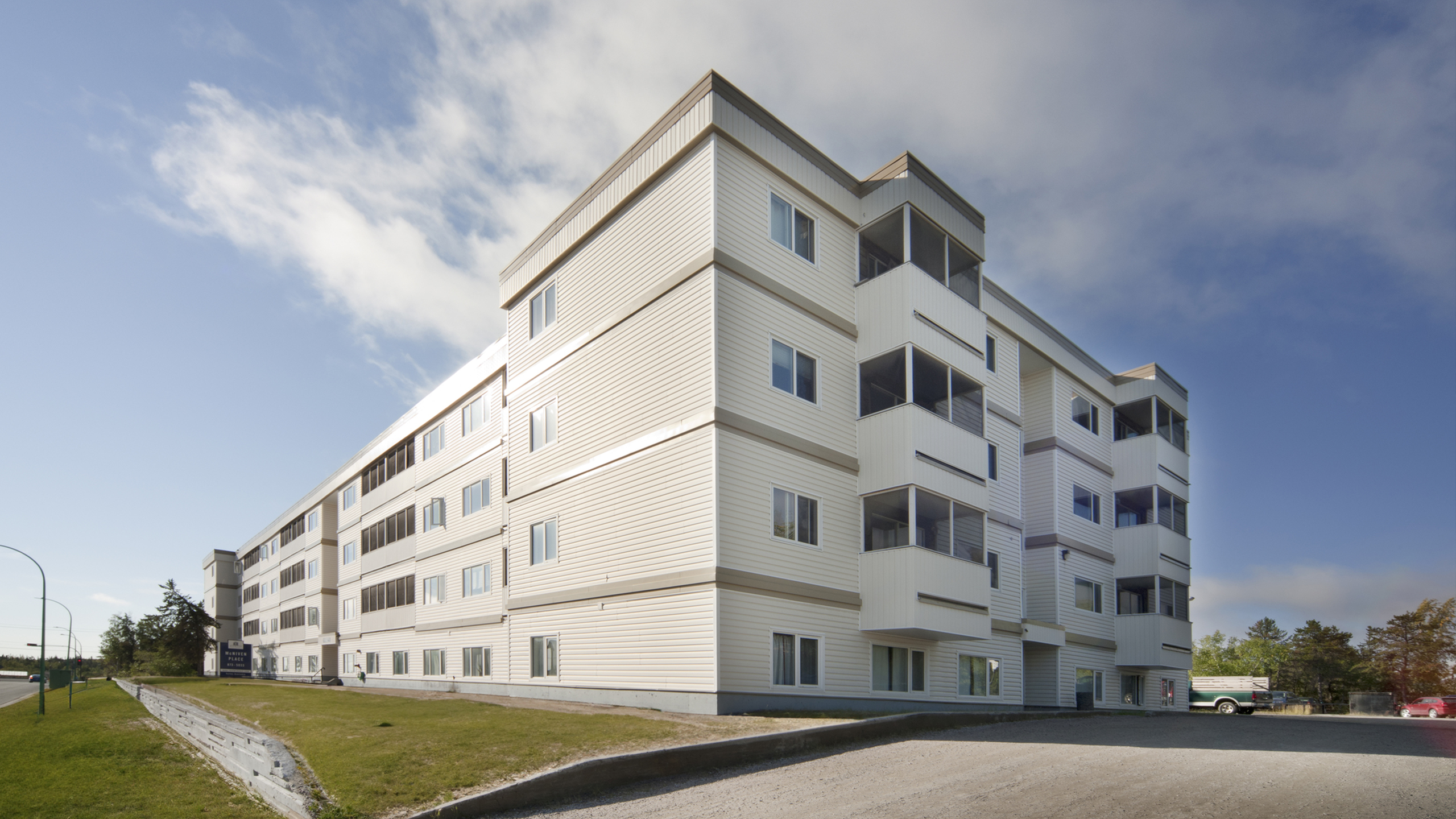 Bright and modern multi-story apartment building with large windows, surrounded by lush greenery and a clear blue sky.