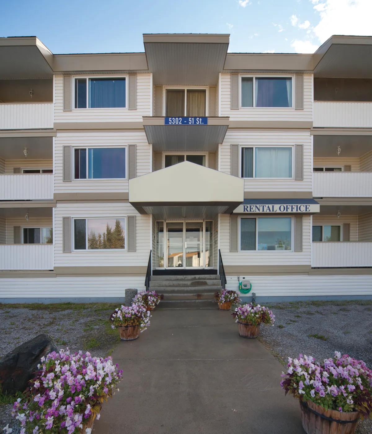 Charming apartment building with welcoming entrance, vibrant flower arrangements, and rental office sign, located on a serene street.