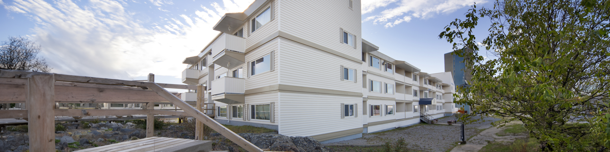 Modern apartment building exterior with clean lines, white siding, and spacious balconies, surrounded by lush greenery under a bright blue sky.
