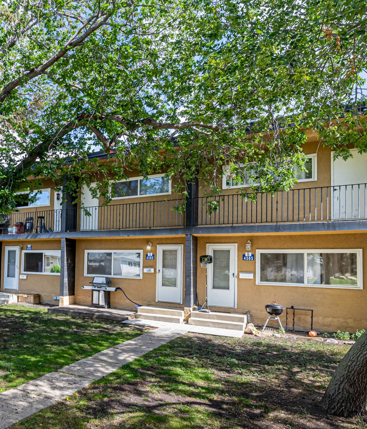 Charming multi-unit building with a picturesque lawn, shaded by trees and featuring a welcoming exterior for a cozy living experience.