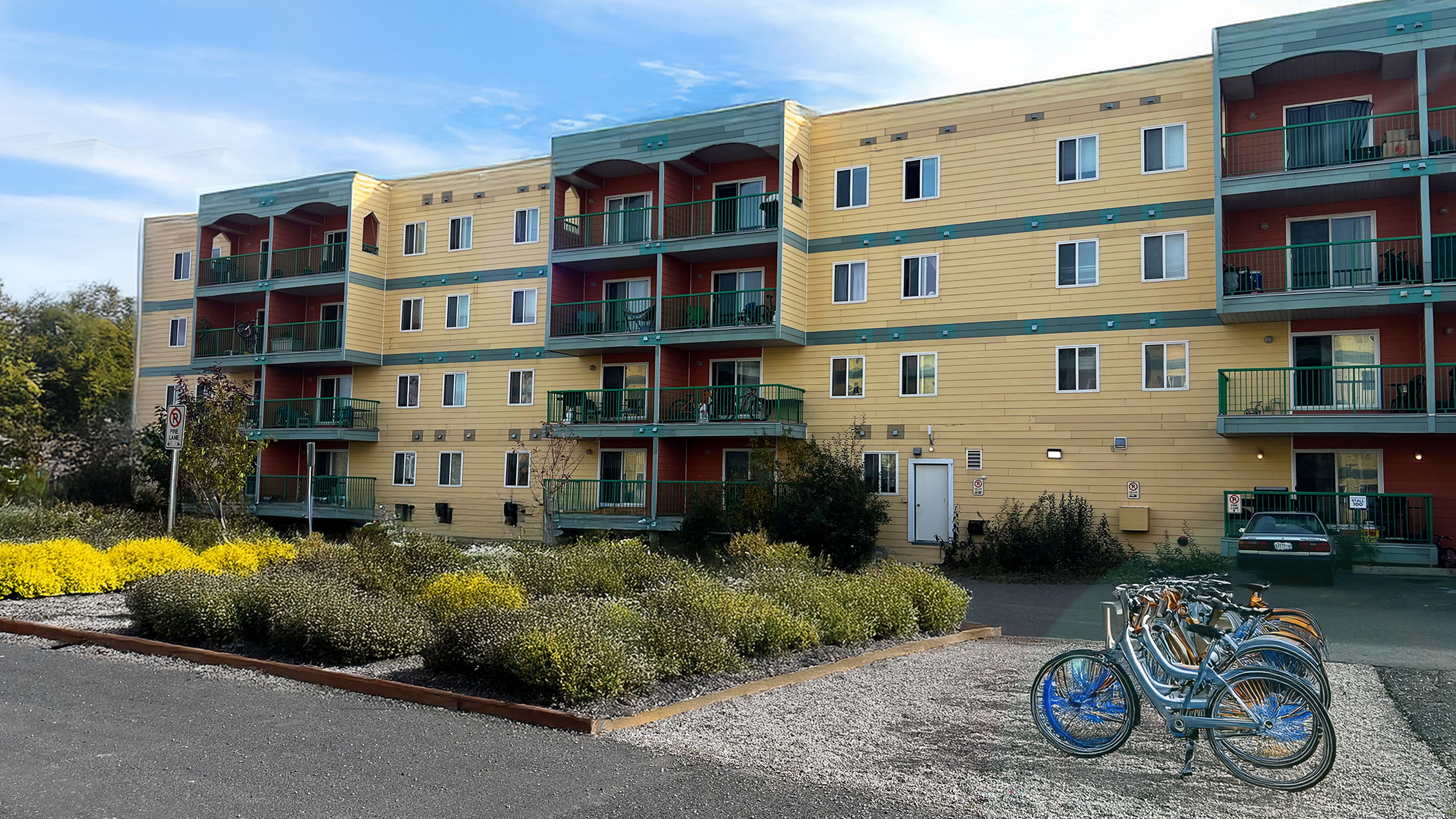 Charming four-story apartment building with vibrant yellow siding, surrounded by lush greenery and bike racks for convenient urban living.