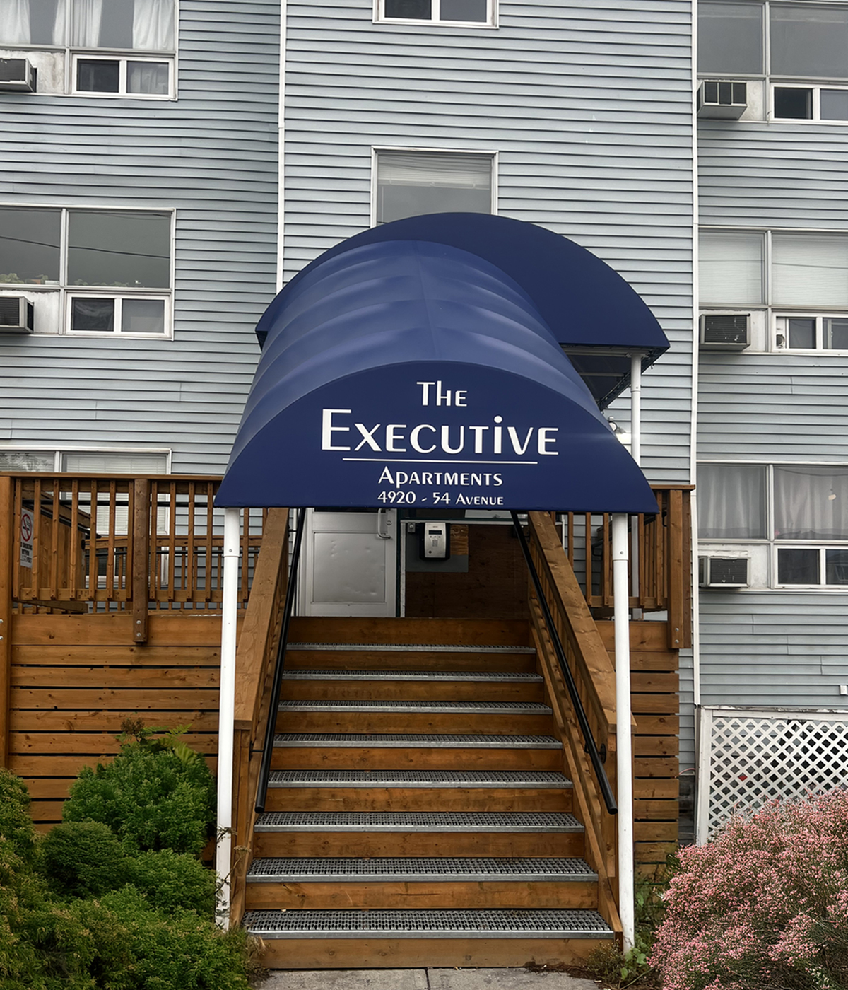 Inviting entrance of The Executive Apartments at 4920 54 Avenue, featuring a stylish navy canopy and well-maintained wooden steps.