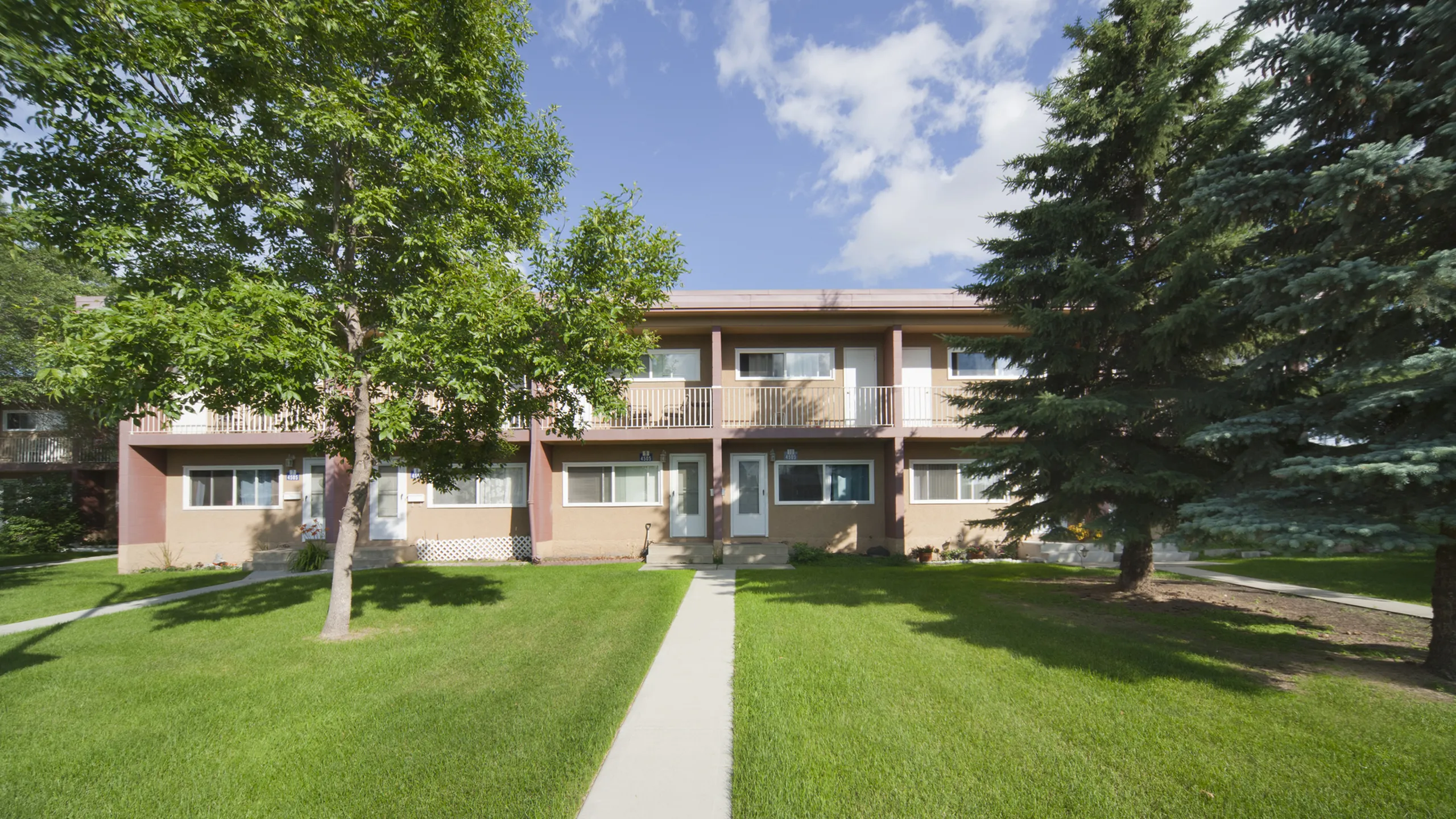 Charming two-story apartment building with manicured lawn, lush trees, and inviting balconies under a bright blue sky.
