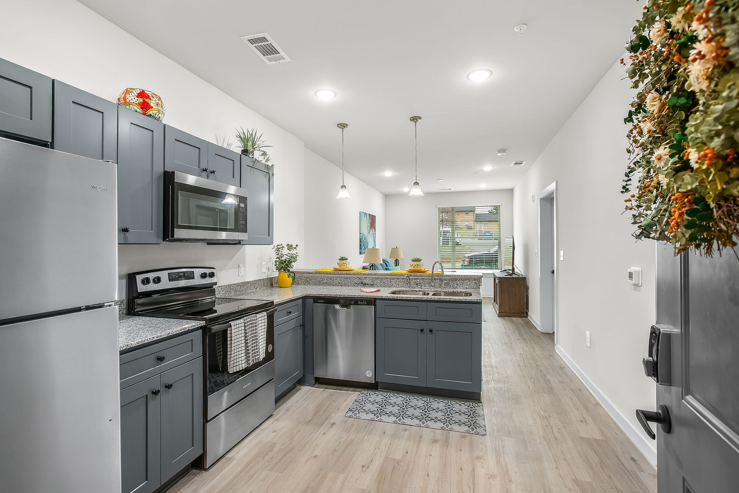 Modern open-concept kitchen with sleek gray cabinets, stainless steel appliances, and elegant pendant lighting, leading to a bright living area with large windows.