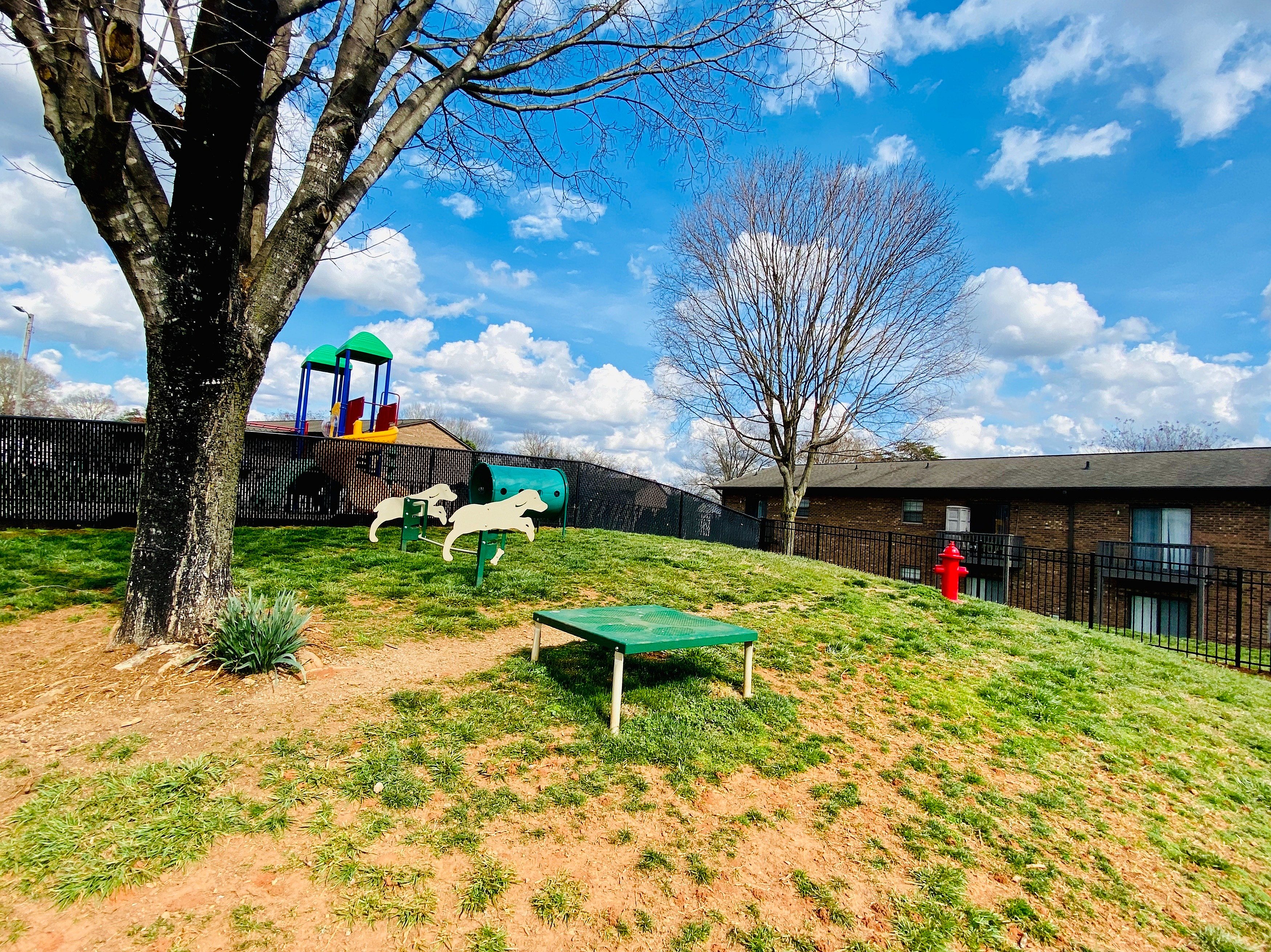 Inviting apartment community with a vibrant outdoor area featuring a playful playground, lush green lawns, and charming park benches set against a clear blue sky.