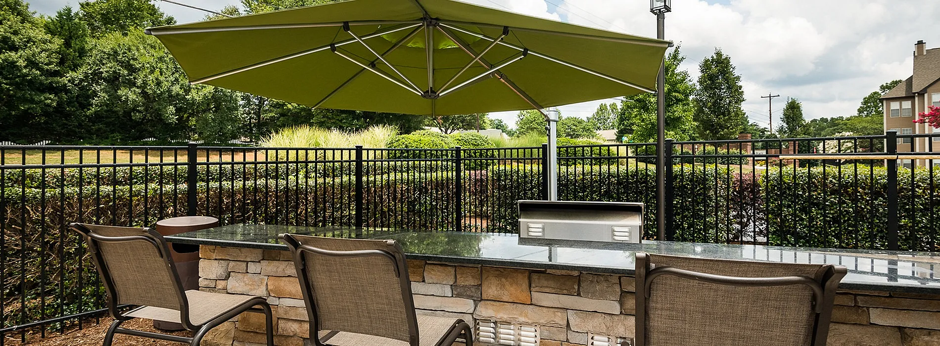 Charming outdoor patio with modern bar seating and sleek stone counter under a vibrant green umbrella, offering a relaxing view of lush greenery.