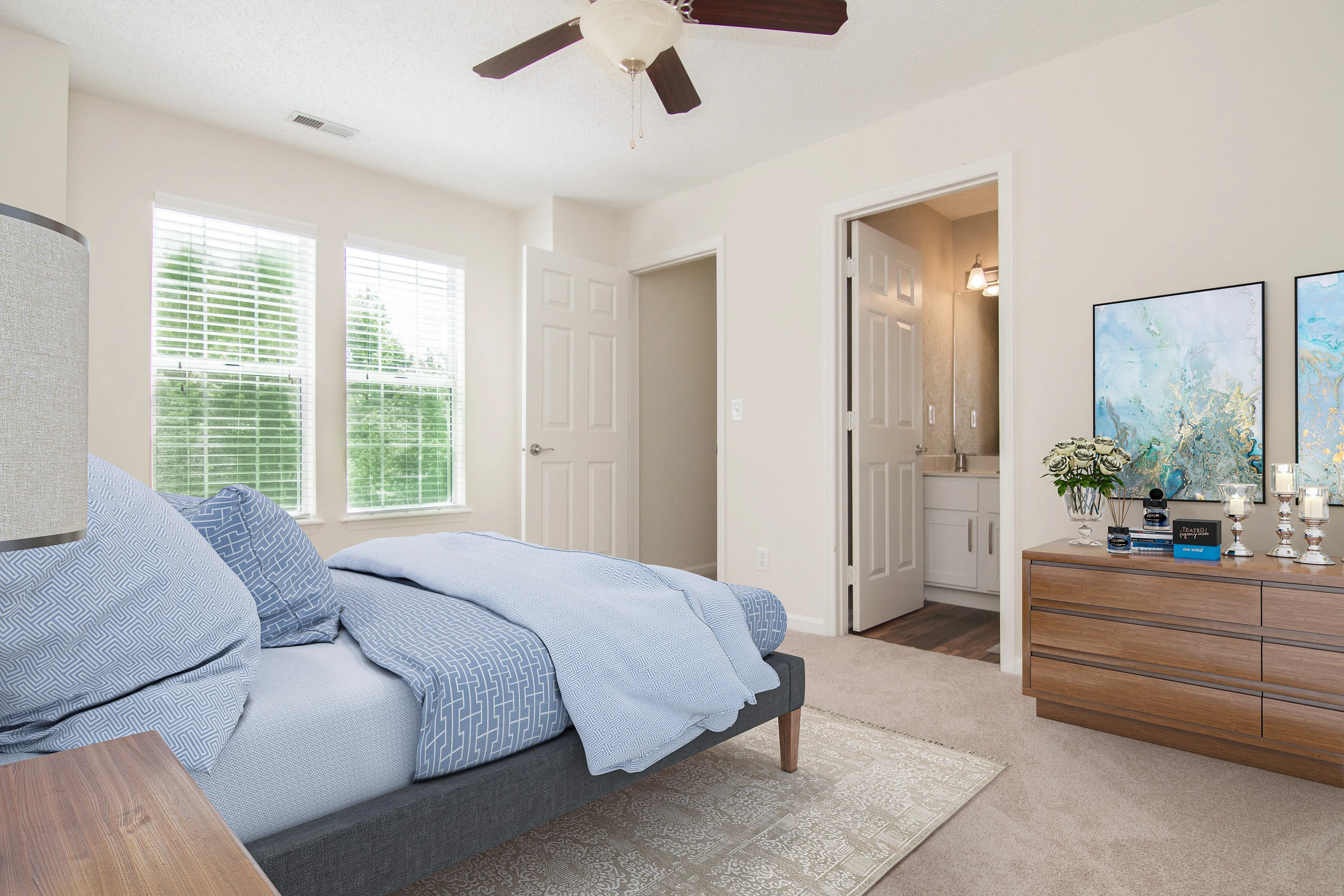 Bright and inviting bedroom with a cozy blue bed, elegant dresser, and soft carpet, enhanced by natural light from large windows.