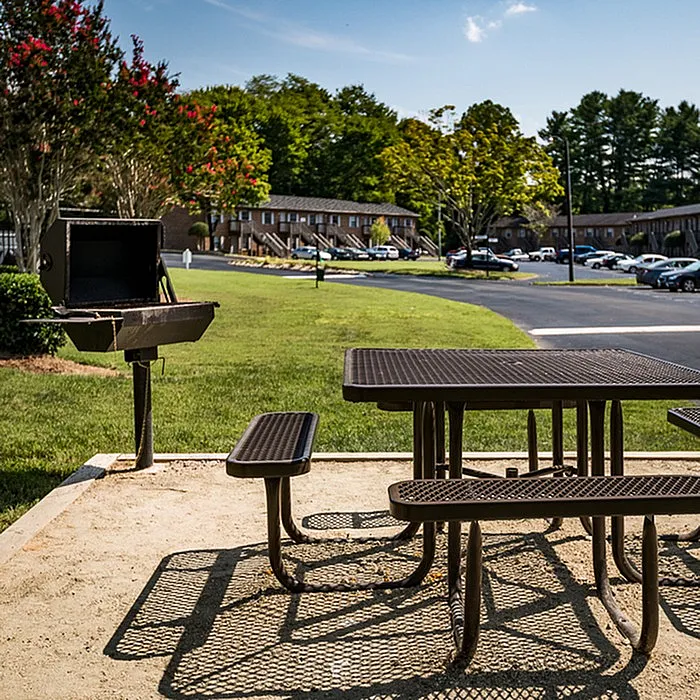 Charming outdoor area featuring a picnic table and grill, perfect for gatherings, set against a backdrop of lush greenery and apartment buildings.