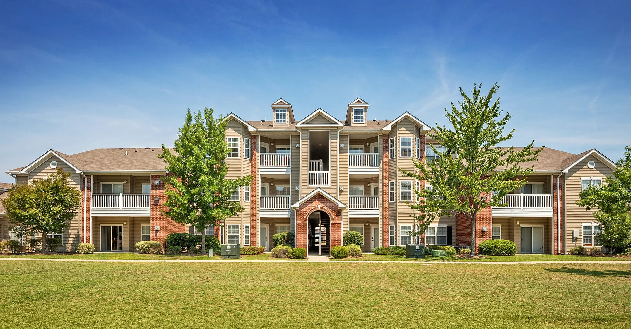 Charming mid-rise apartment building with elegant balconies, surrounded by lush green lawns and trees, perfect for relaxed living.