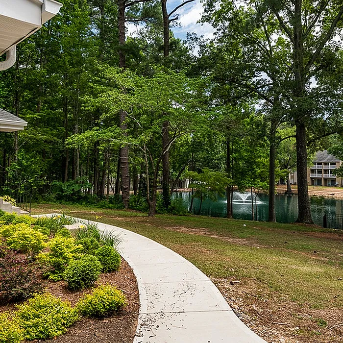Charming garden pathway leading to serene lake with water fountain amidst lush greenery.