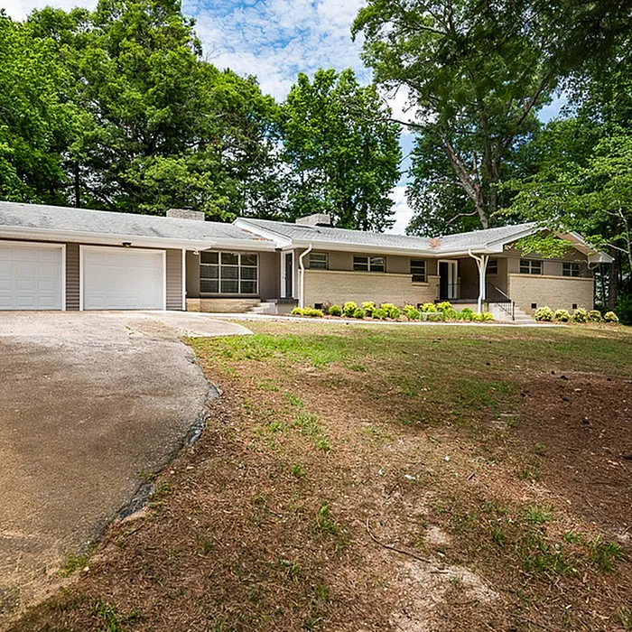 Charming mid-century ranch-style home with a spacious driveway and lush greenery surrounding.