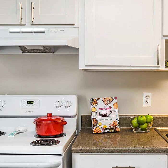 Inviting kitchen corner with sleek countertops, modern stove, and chic white cabinetry. A vibrant red pot adds a pop of color, while a bowl of fresh green apples and a cookbook bring a homely touch.
