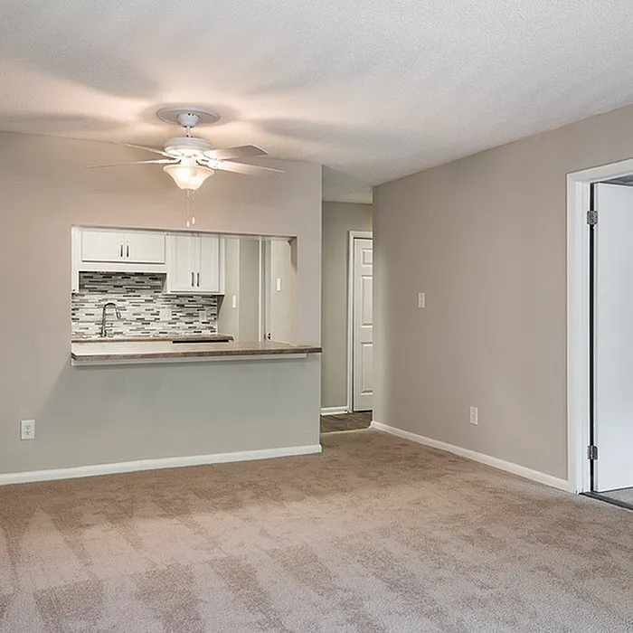 Modern apartment interior showcasing a cozy open-concept living area with beige carpet, sleek kitchen with stylish backsplash, and a ceiling fan for comfort.