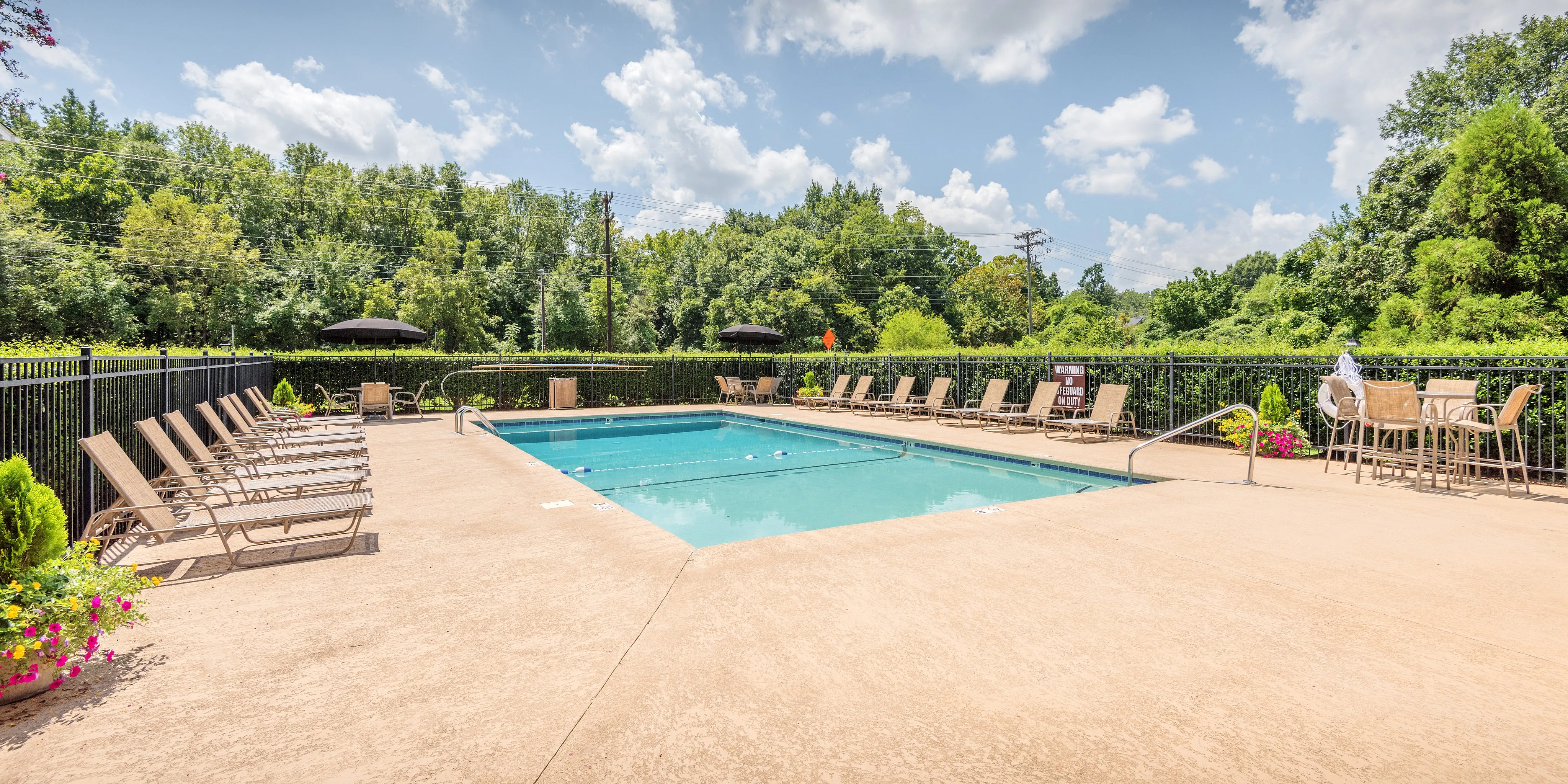 Inviting outdoor pool area with lounge chairs and seating, surrounded by lush greenery under a bright blue sky.