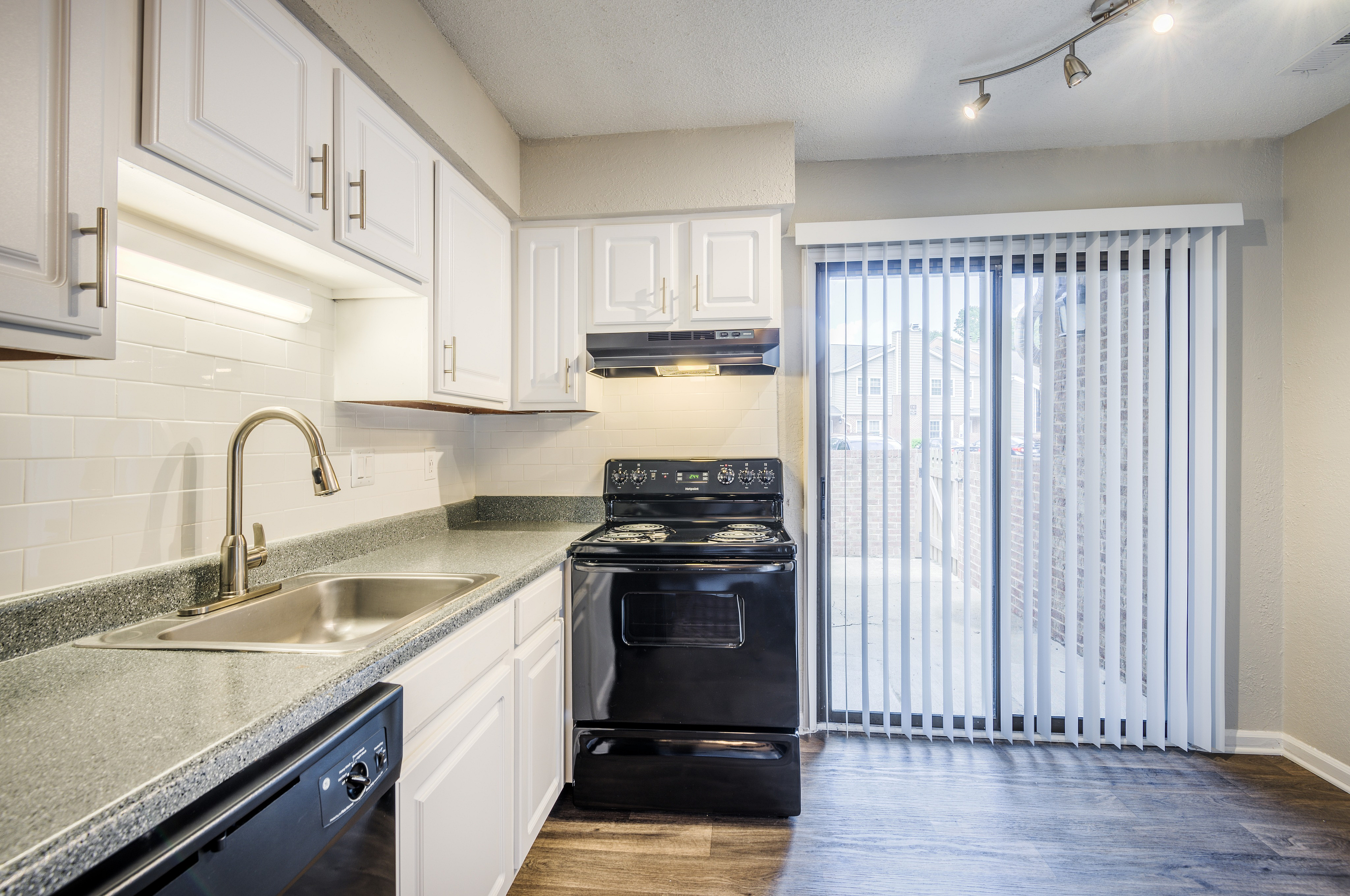 Modern kitchen with sleek white cabinets, granite countertops, and stainless steel fixtures, opening to a sunlit patio through elegant vertical blinds.