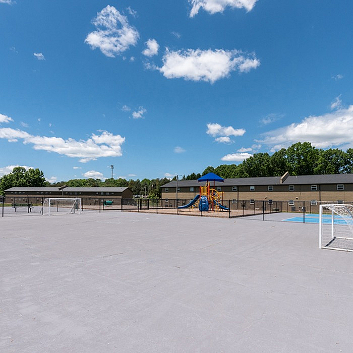 Sunlit outdoor playground and sports court in a serene residential community, surrounded by lush greenery and modern buildings.