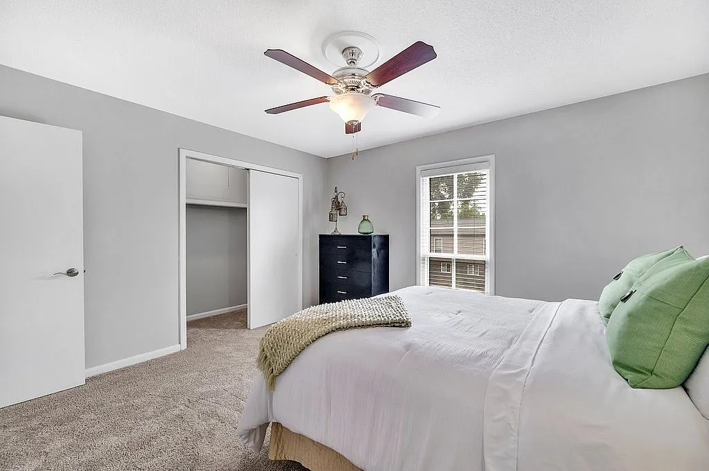 Bright and welcoming bedroom with soft neutral walls, featuring a cozy bed adorned with green pillows, a modern ceiling fan, and ample natural light streaming through a large window.