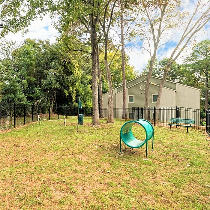 Charming fenced dog park with agility tunnel, surrounded by lush greenery near a cozy residential building.