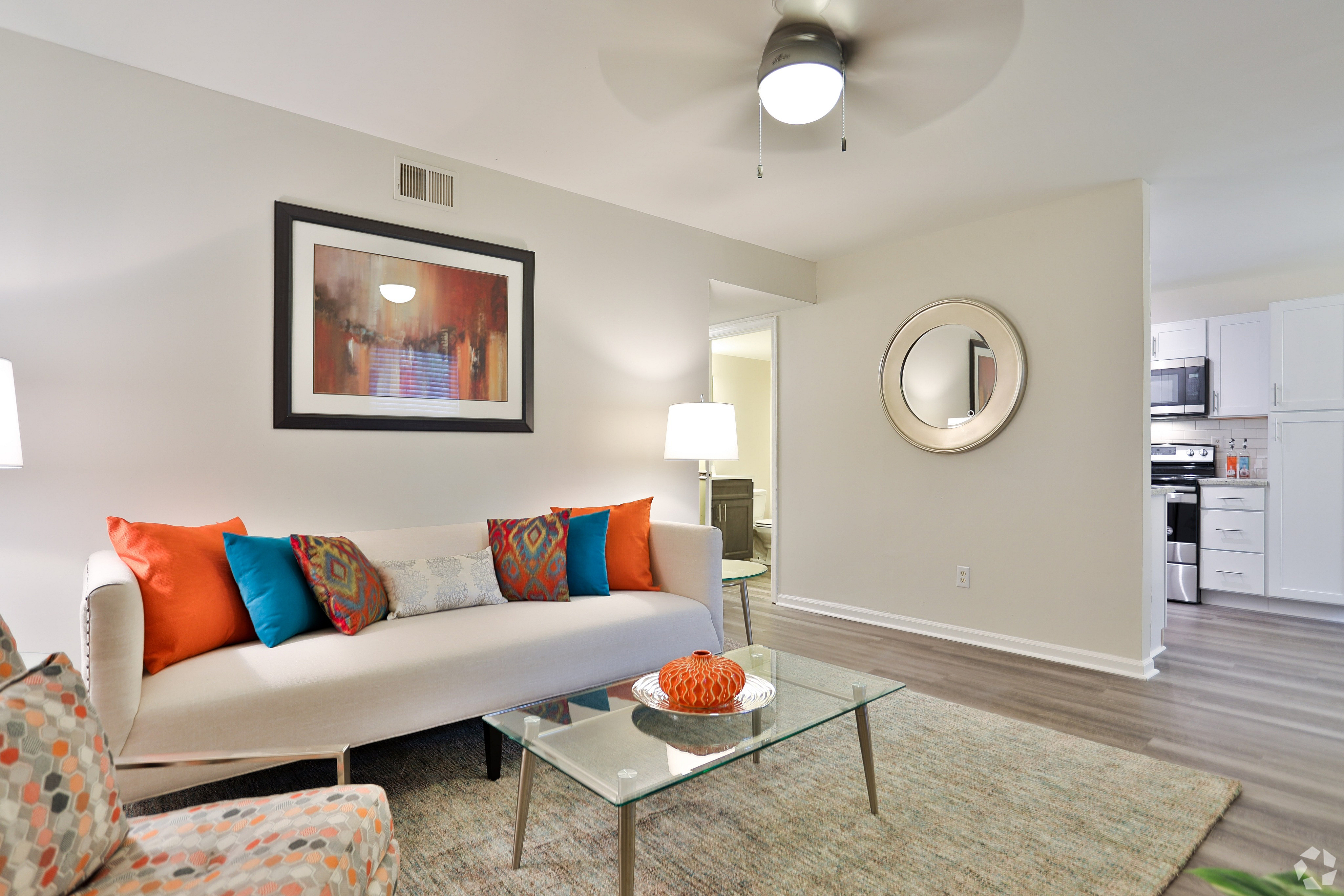 Elegant living room featuring a chic white sofa with vibrant, colorful pillows, a glass coffee table with decorative vase, and stylish artwork. Modern open-concept design with a glimpse into a sleek kitchen.