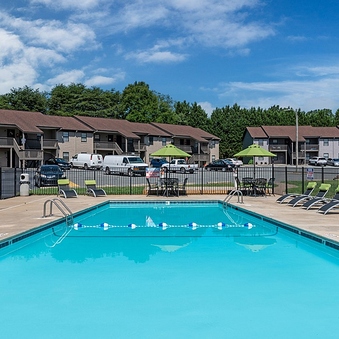 Inviting apartment complex featuring a sparkling swimming pool, surrounded by lounge chairs and vibrant green umbrellas, under a clear blue sky.