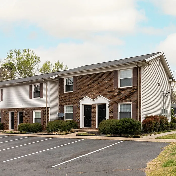 Charming two-story apartment building with brick facade, white siding, and well-maintained landscaping, featuring ample parking space and surrounded by lush greenery.
