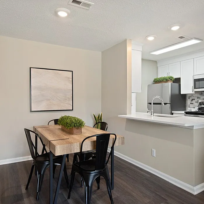 Modern apartment dining and kitchen area featuring a sleek wooden table with black chairs, contemporary white cabinets, and stainless-steel appliances.