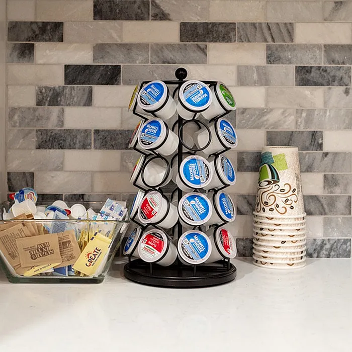 Modern kitchen countertop featuring a stylish coffee pod carousel, decorative paper coffee cups, and a bowl of assorted sweeteners against a chic tile backsplash.