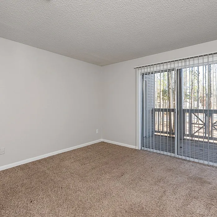 Bright apartment room with plush carpet and large sliding glass doors opening to a serene private balcony.