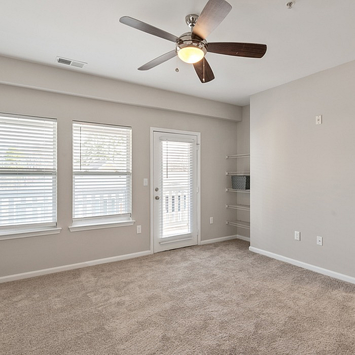Bright and airy spacious room with plush carpet, featuring large windows, a ceiling fan, and a door leading to a private balcony.