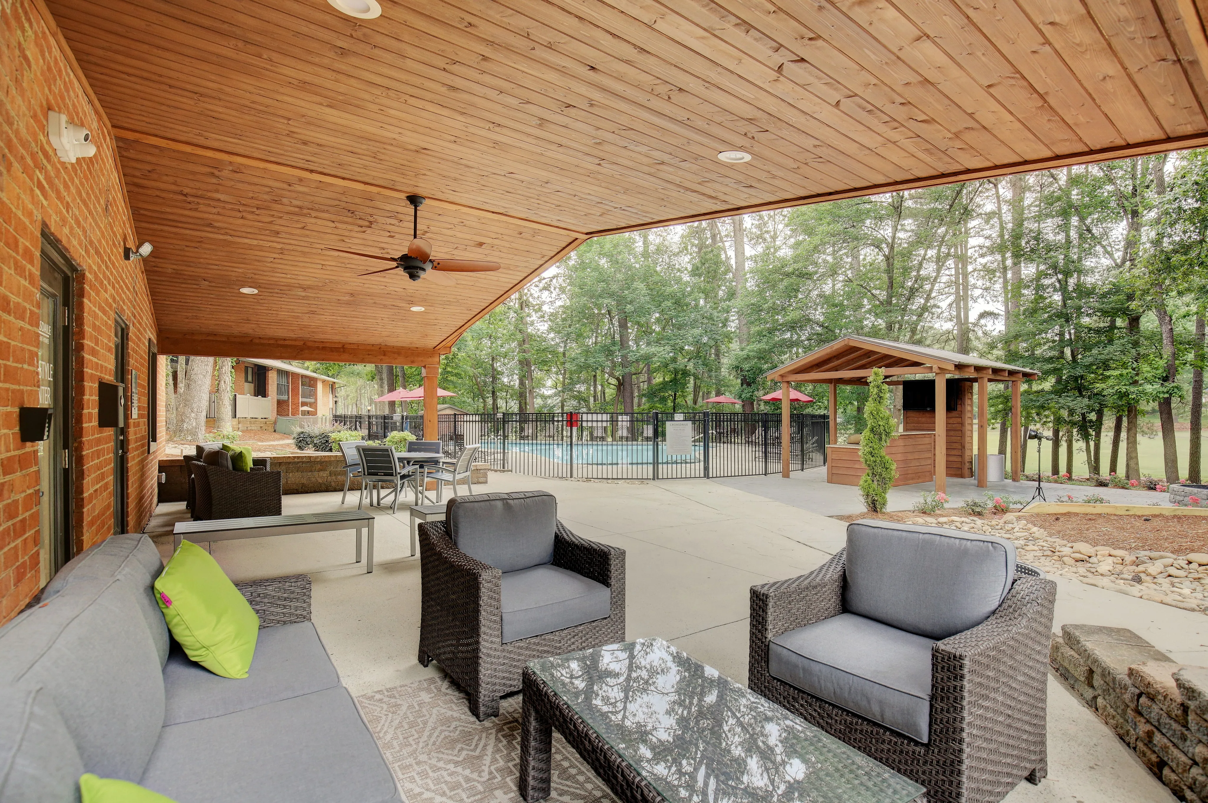 Inviting outdoor patio with cozy seating, wood-paneled ceiling, and views of a sparkling pool surrounded by lush trees.