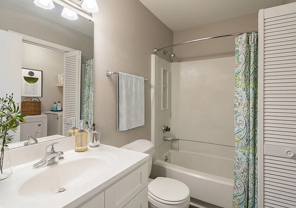 Bright and modern bathroom featuring sleek white fixtures, a decorative shower curtain, and a glimpse of a cozy laundry area in the background.