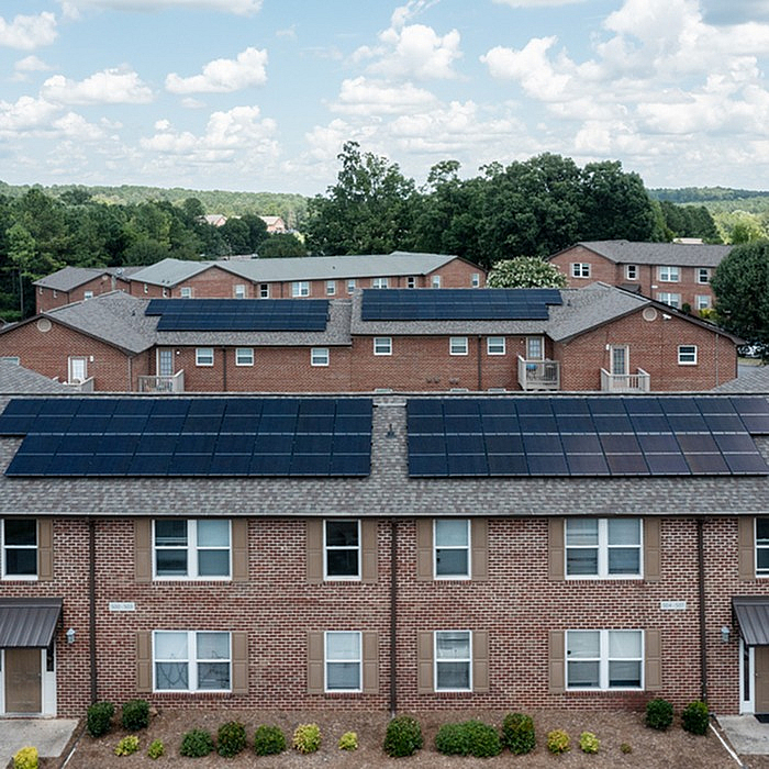 Charming brick apartment complex with solar panels on rooftops set against lush green landscape and blue sky.