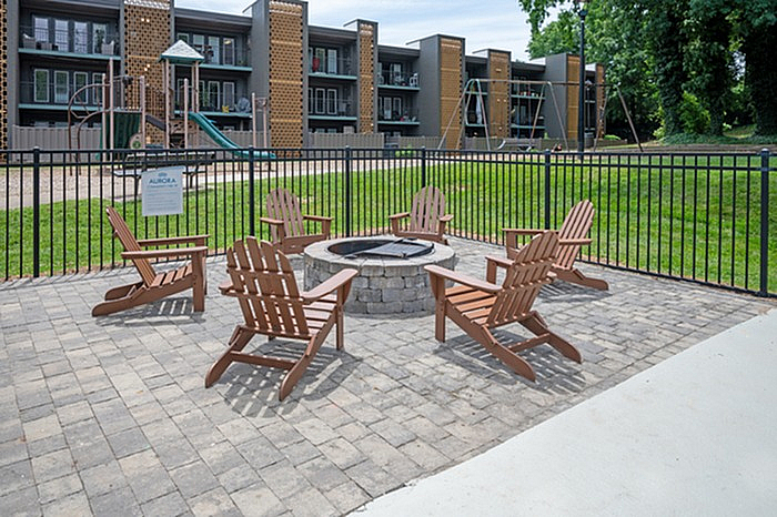 Charming outdoor patio with cozy Adirondack chairs surrounding a fire pit, set against modern apartment buildings and a lush green lawn.