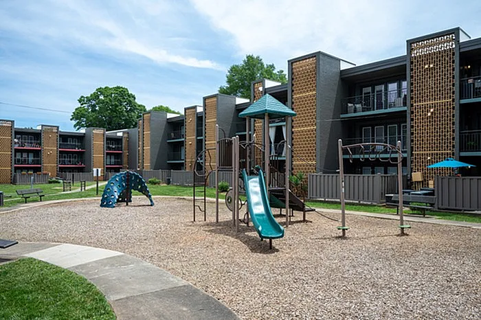 Modern apartment complex with children's playground featuring slides and climbing structures, surrounded by green spaces and patio seating.