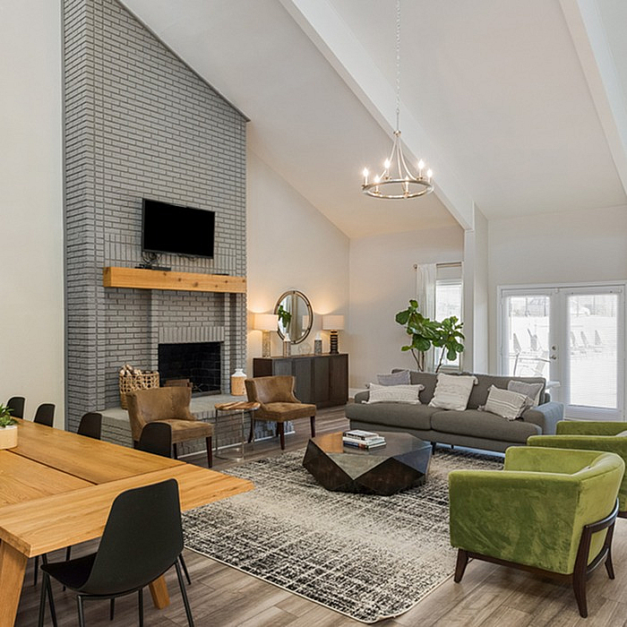 Elegant living room with high ceilings, modern chandelier, and cozy seating area featuring a stylish gray sofa and vibrant green chairs. The space includes a classic brick fireplace, large windows, and natural light creating a warm, inviting ambiance.