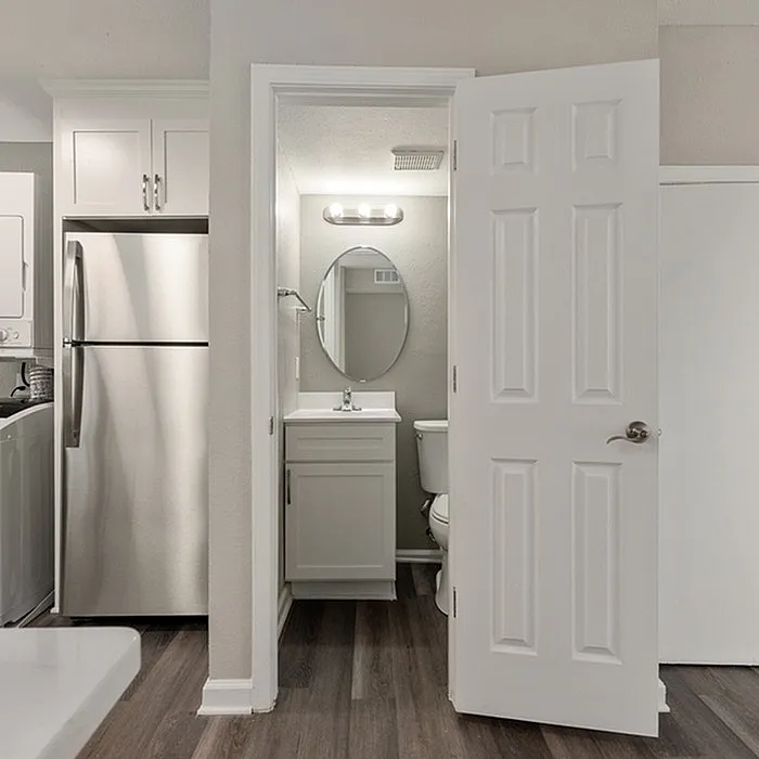 Modern apartment view featuring a sleek stainless steel refrigerator next to a compact, stylish bathroom with elegant lighting and an oval mirror.