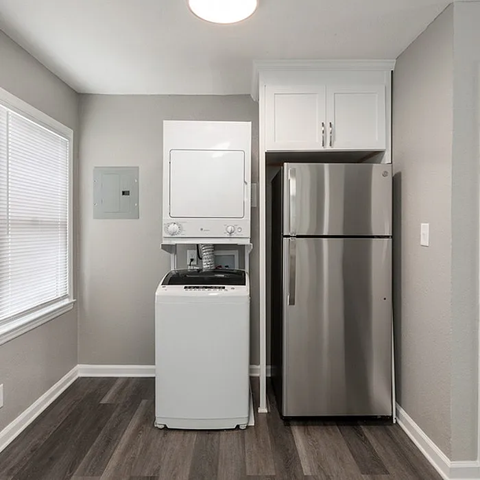 Modern laundry area with a sleek stackable washer-dryer combo next to a stainless steel refrigerator, accented by stylish cabinetry and elegant flooring.