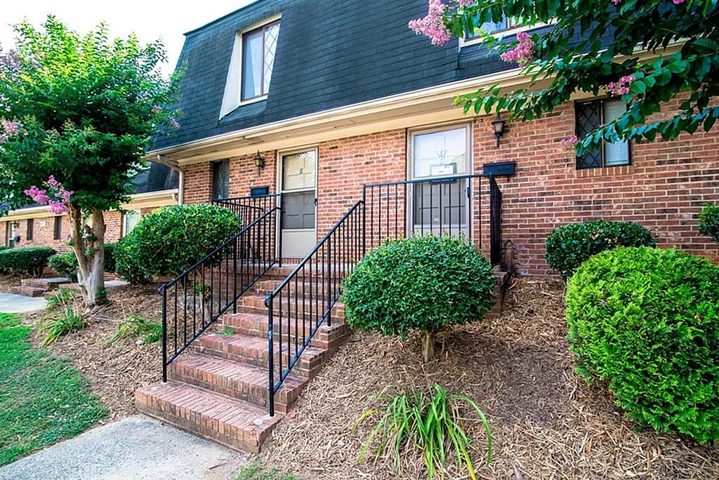 Charming brick townhouse with welcoming staircase, surrounded by lush greenery and vibrant flowering trees.