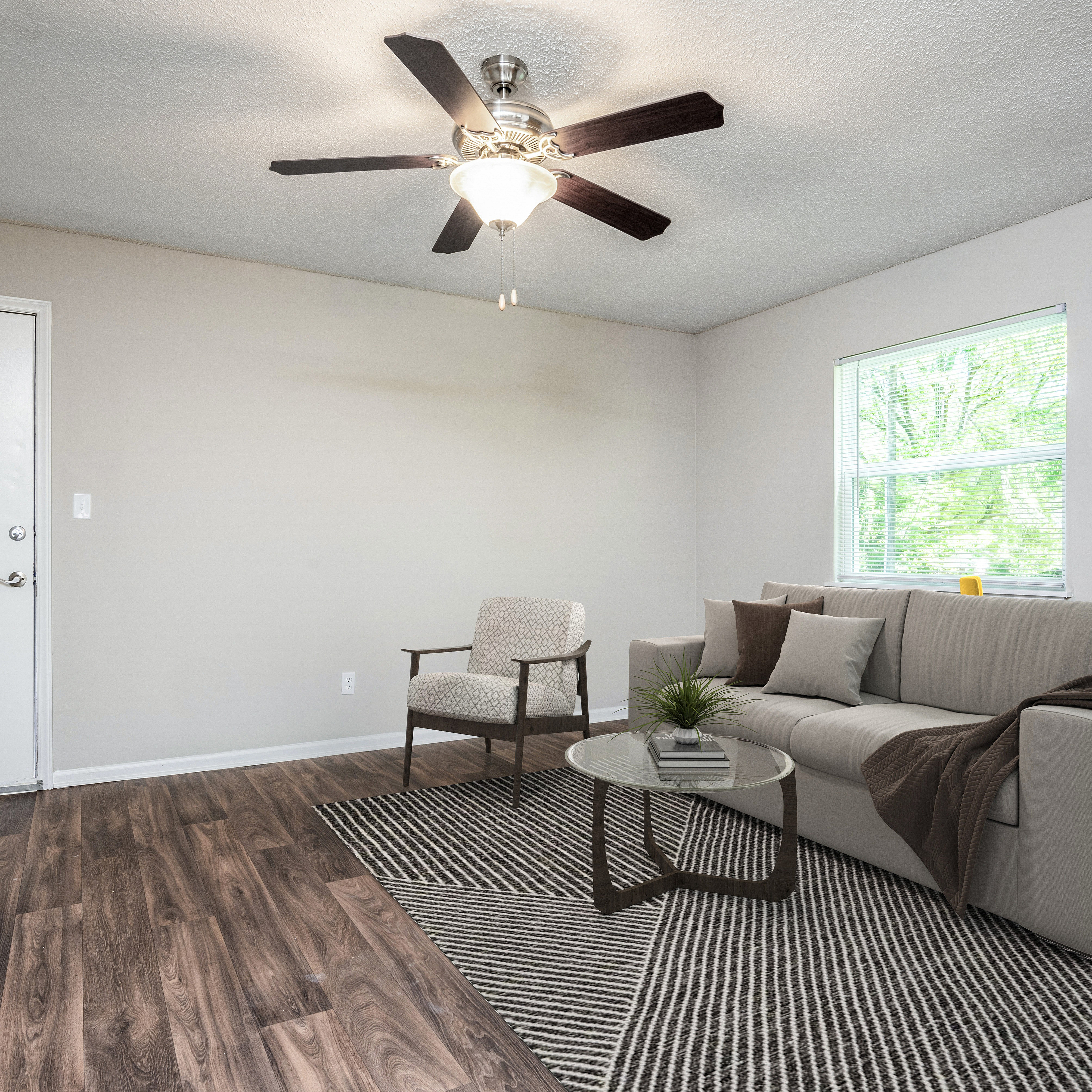 Bright and inviting living room featuring a modern gray sofa, elegant armchair, and stylish ceiling fan, complemented by a chic striped rug and a leafy view outside the window.