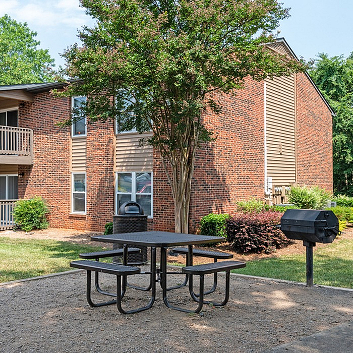 Charming apartment exterior with a cozy brick facade, complemented by a lush tree and a convenient outdoor picnic area with a barbecue grill.