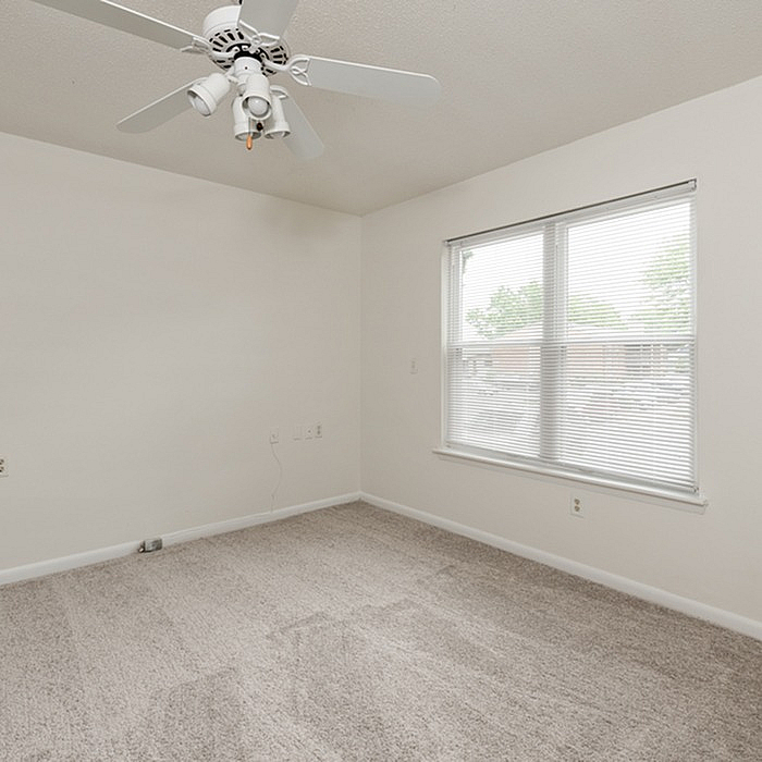 Bright, airy bedroom with large window and ceiling fan, featuring plush carpeting and a neutral color palette for a cozy ambiance.