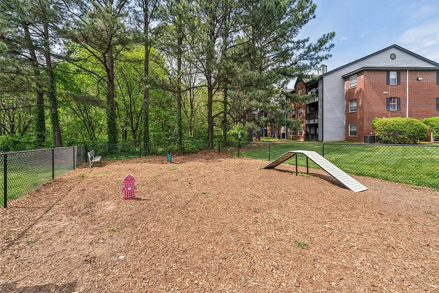 Charming dog park with agility ramp and vibrant greenery, adjacent to modern brick apartment building.