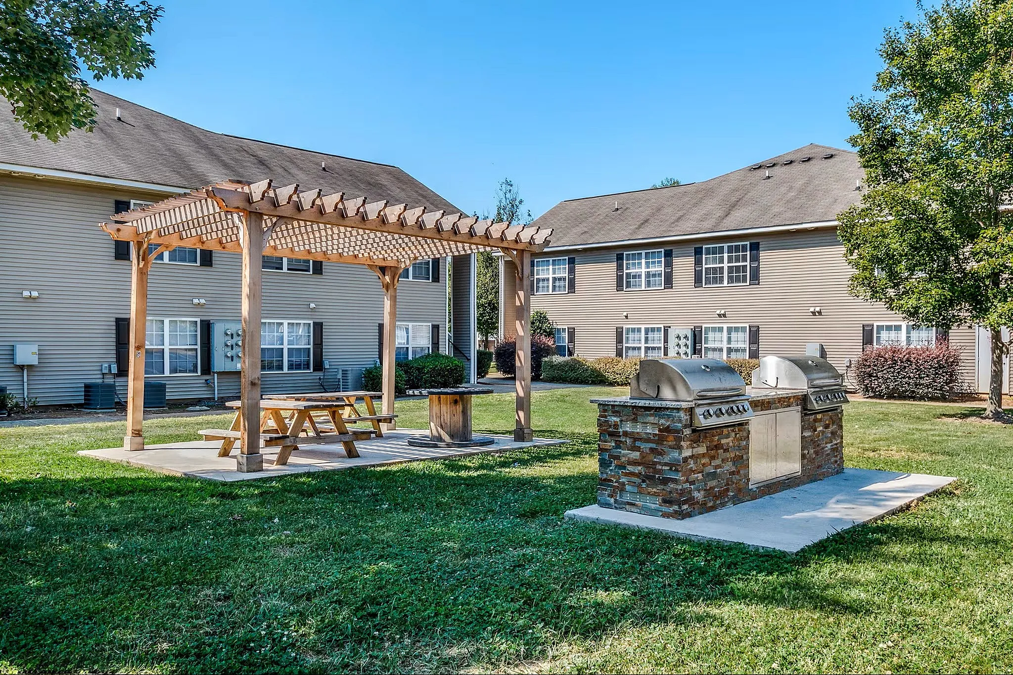 Inviting outdoor community space featuring a charming wooden pergola, picnic table, and modern grilling station set against stylish apartment buildings.